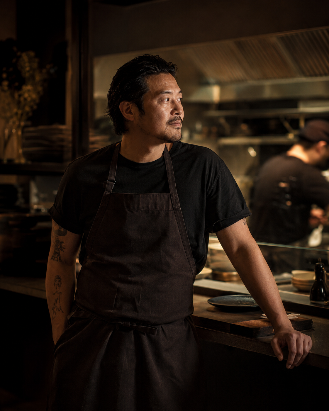 Mika Tan, chef and co-founder of Ember House, near the pass in the kitchen — relaxed, looking away from the camera, lit by warm side light.