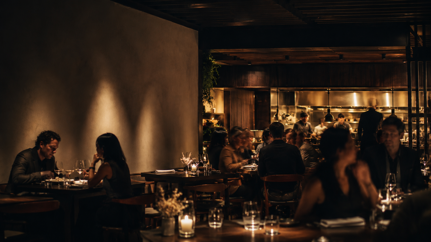The Ember House dining room at night — warm pools of light over dark timber, diners in natural movement, the open kitchen glowing at the back.