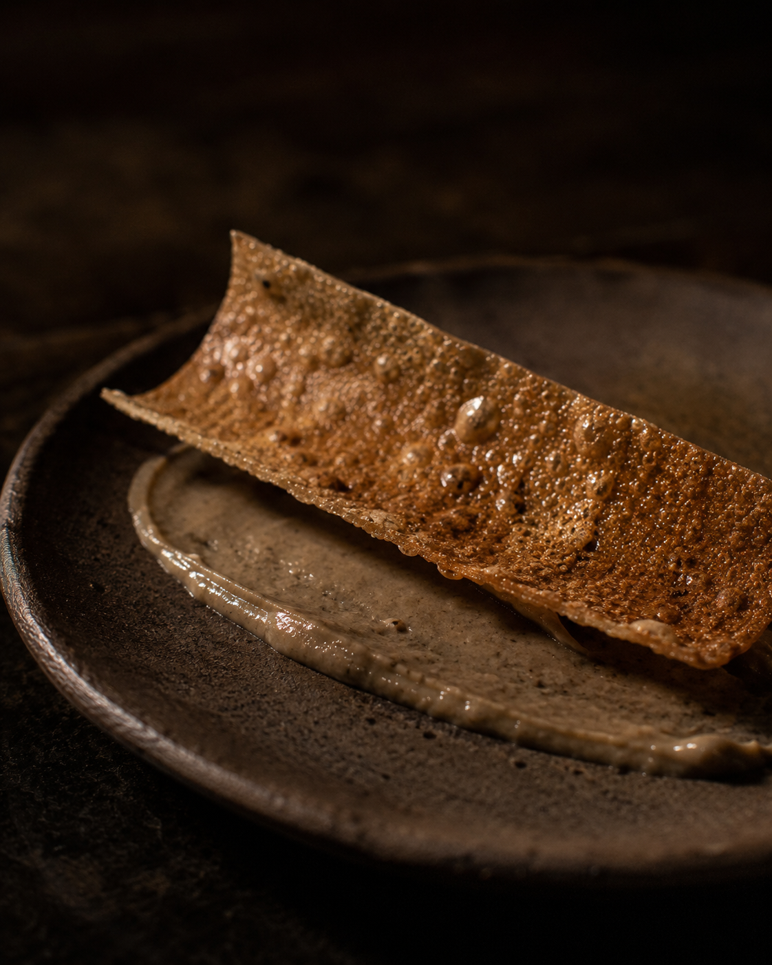 Crisp duck-skin cracker resting over a smear of smoked eggplant on dark ceramic.