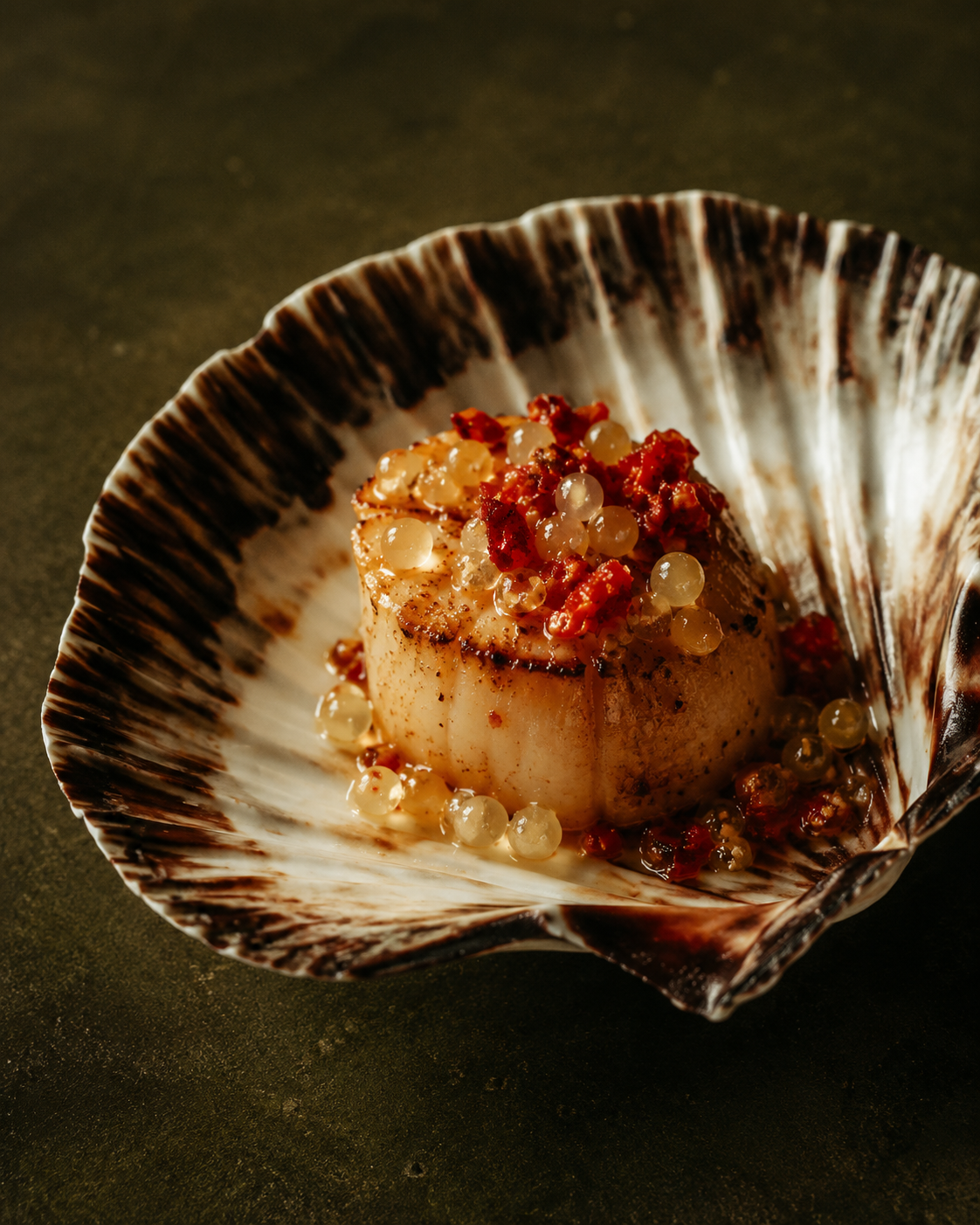 Single seared scallop in its shell, dressed with fermented chilli and beads of finger lime, shot close and warm.