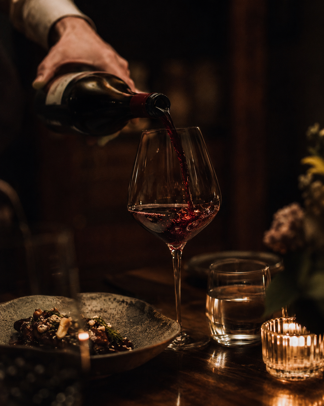 Glass of wine being poured at the table, deep colour against dark timber.